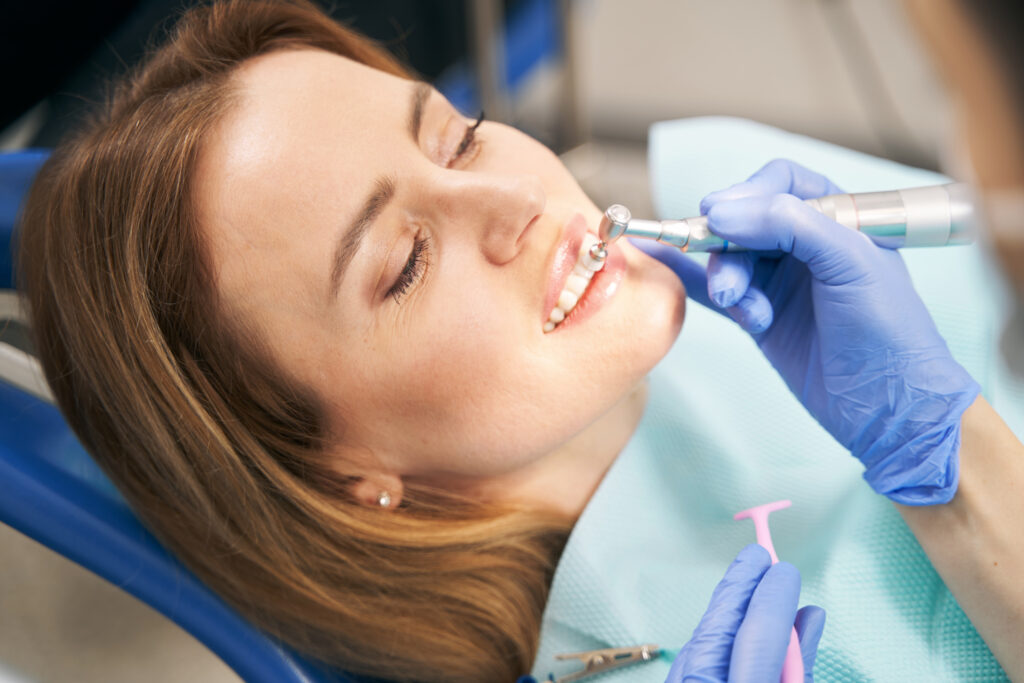 Close up of doctor in sterile gloves cleaning and polishing female teeth with dental polisher while woman lying in dental chair